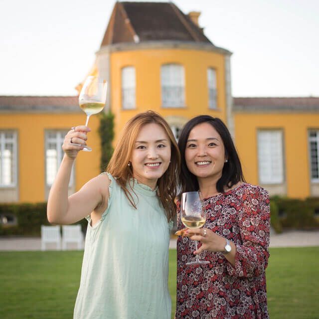 Two women toast with wine glasses, smiling in front of a sunny yellow castle, enjoying a joyful moment.